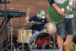 Chris Correia (large white male in a black t-shirt and turquoise hat, seated behind a drumset) and Thom Dunn(tall white male, mostly shown here as a green semi-hollow electric guitar) of the Roland High Life performing at the Emerson College Uncommon Stage in Boston, Massachusetts